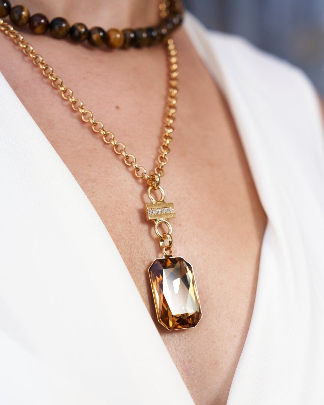 A close-up of a gold chain necklace with a large faceted amber-brown rectangular pendant, worn on a model's neck over a white top.