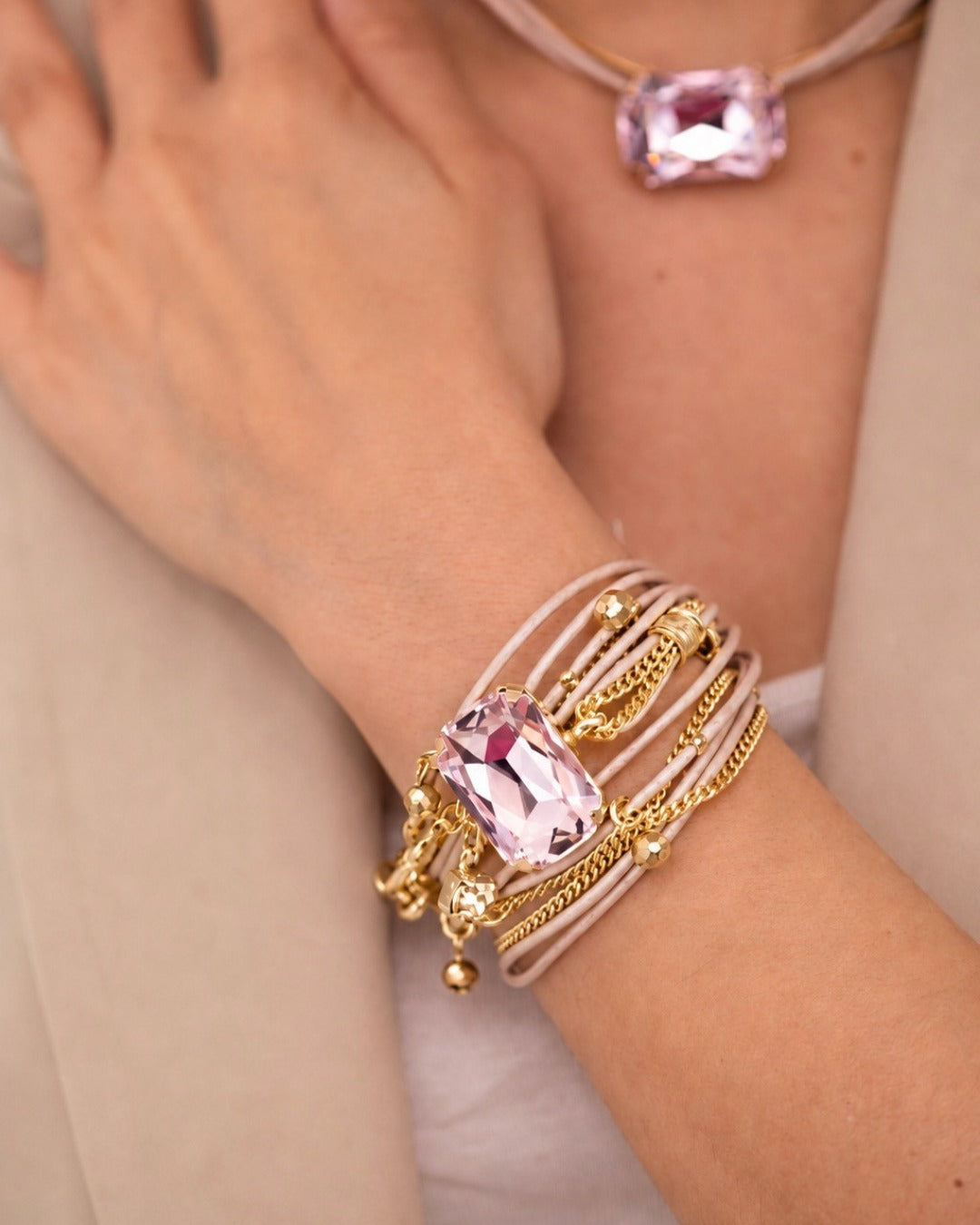 A close-up of a person's wrist wearing a multi-strand bracelet with a large pink gemstone and gold chain details, with a matching pink gemstone necklace partially visible.