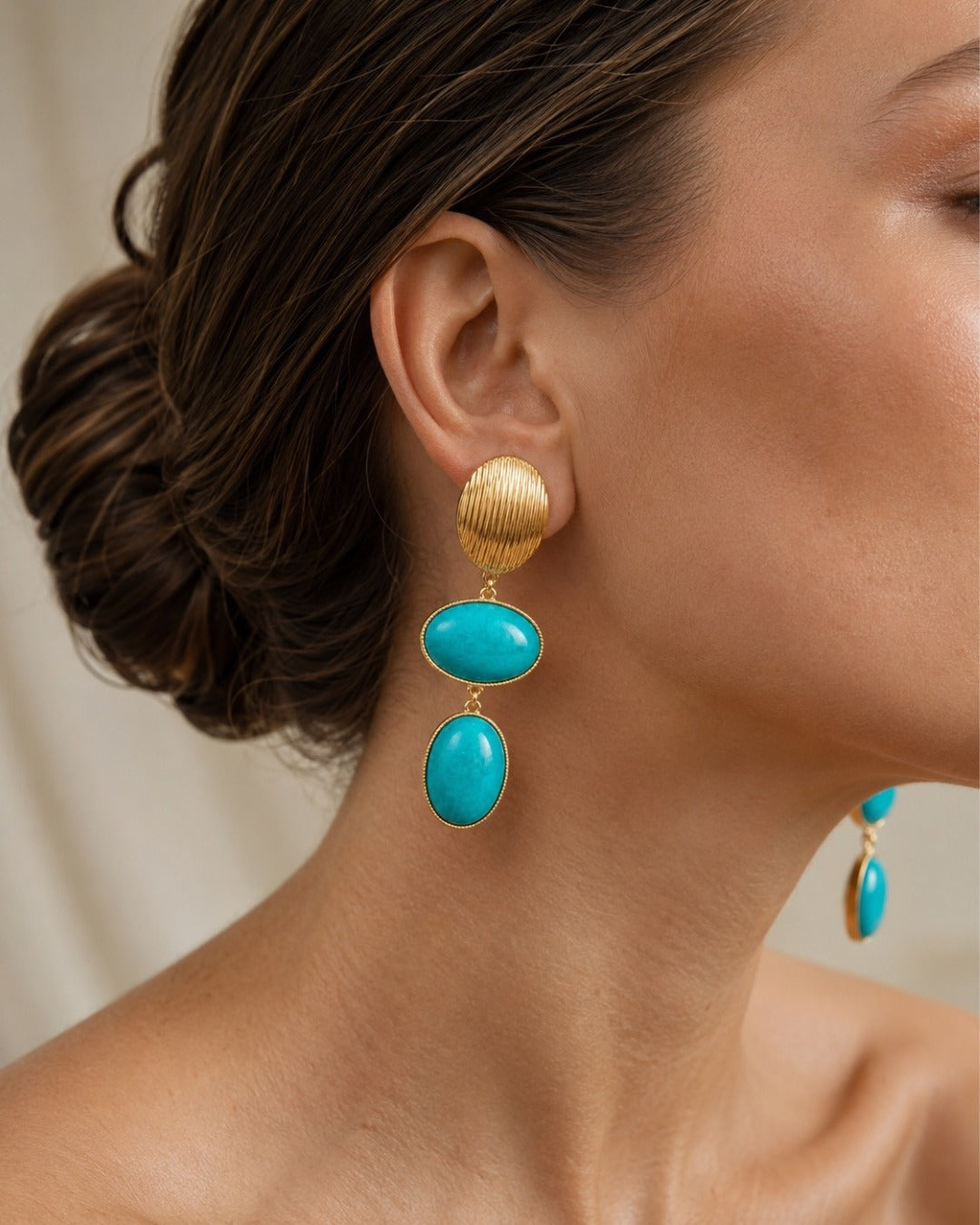 A close-up of a model's ear wearing a gold drop earring with two oval turquoise-blue stones, against a soft beige background.