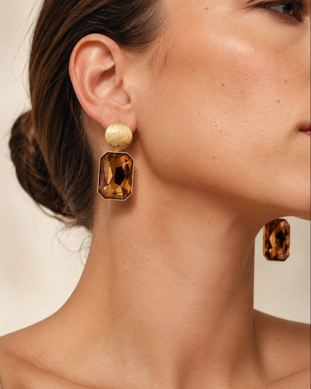 Close-up of a woman wearing a pair of statement drop earrings with textured gold studs and large amber-brown rectangular faceted charms, shown against a soft beige background.