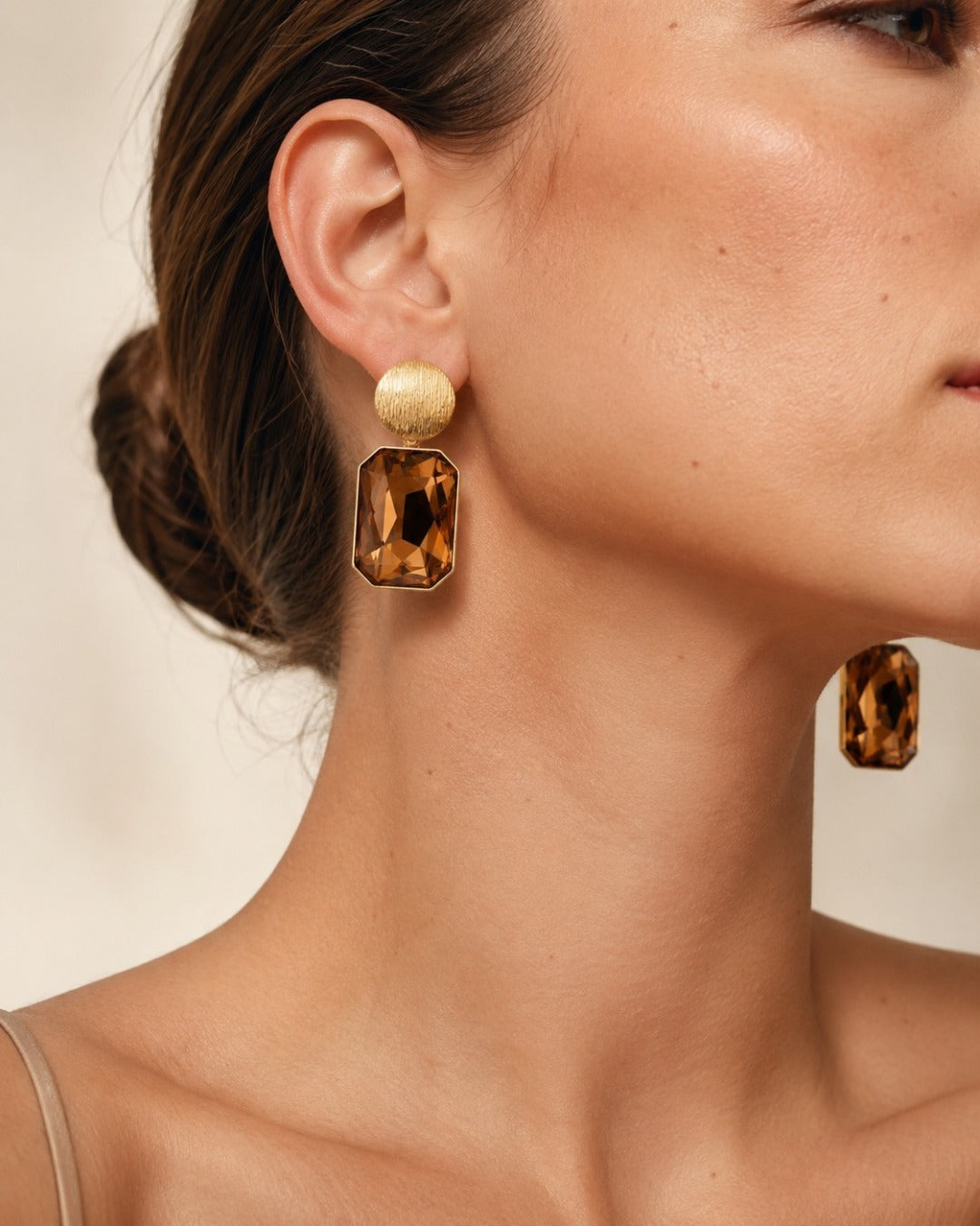 Close-up of a woman wearing a pair of statement drop earrings with textured gold studs and large amber-brown rectangular faceted charms, shown against a soft beige background.