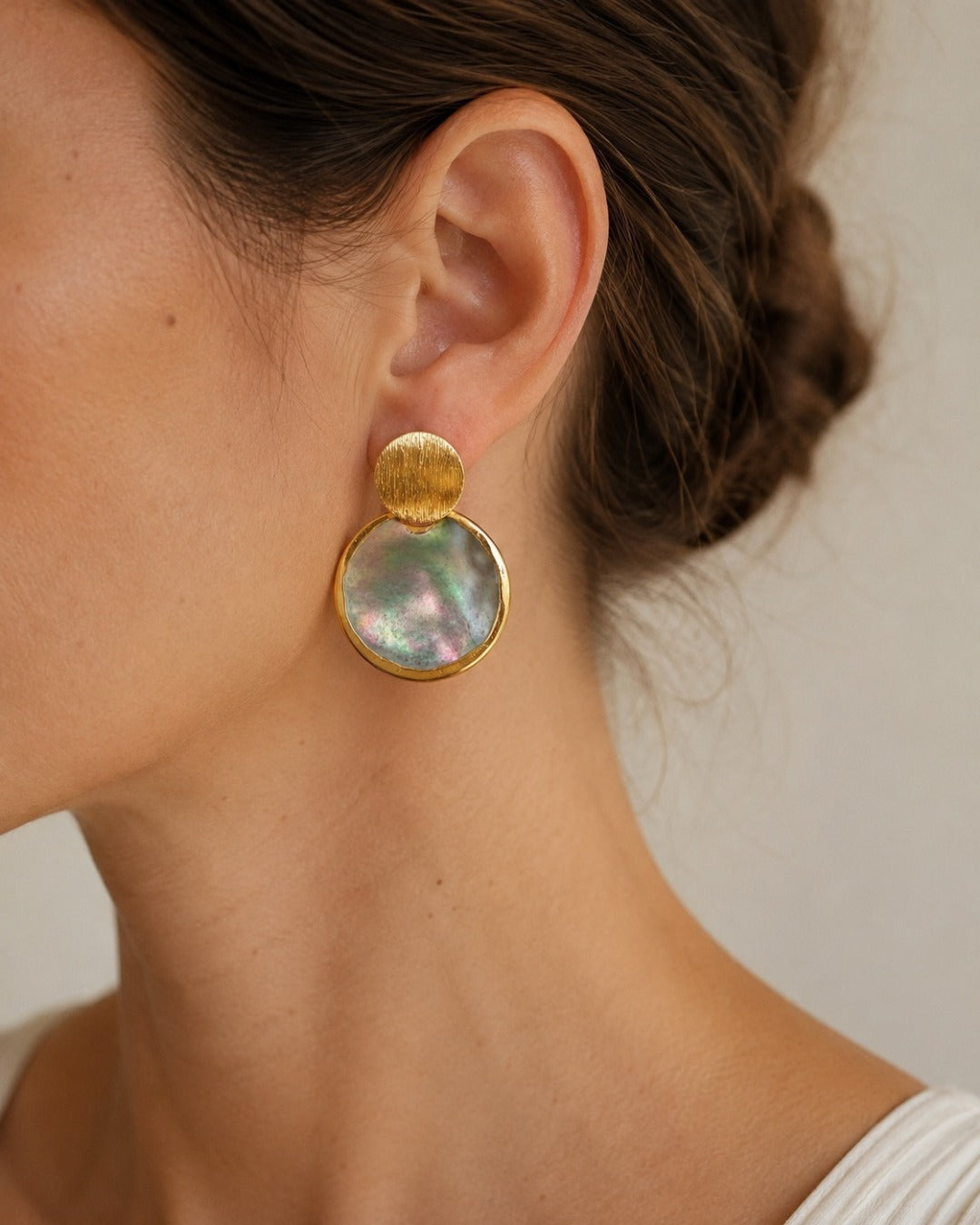 A close-up of a model's ear wearing a gold drop earring with a large, iridescent mother-of-pearl disc.