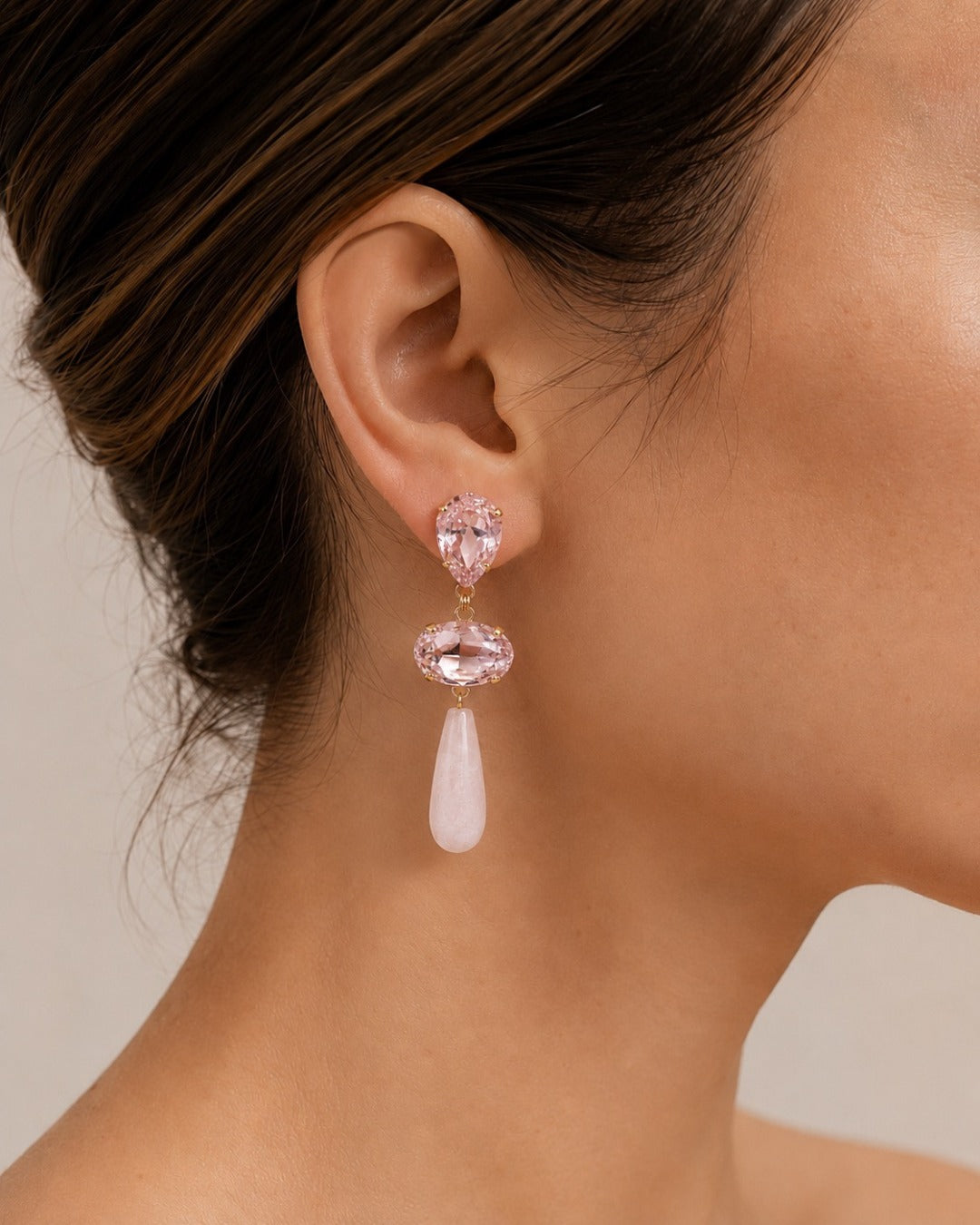 A close-up of a model's ear wearing a dangling earring with two faceted pink gemstones and a teardrop-shaped pale pink stone below.