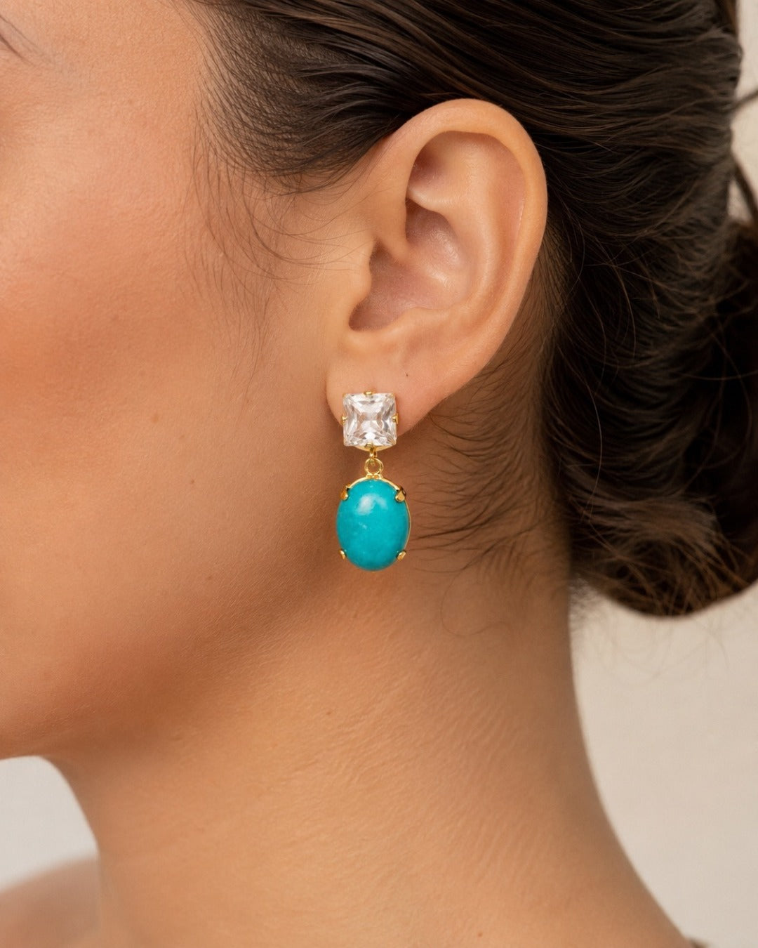 A close-up of a model's ear wearing a gold drop earring with a clear square stud and a dangling oval turquoise stone.