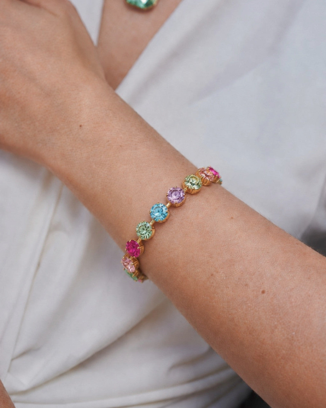 A close-up of a gold bracelet with multiple colorful gemstone flower charms, worn on a person's wrist against a white top.