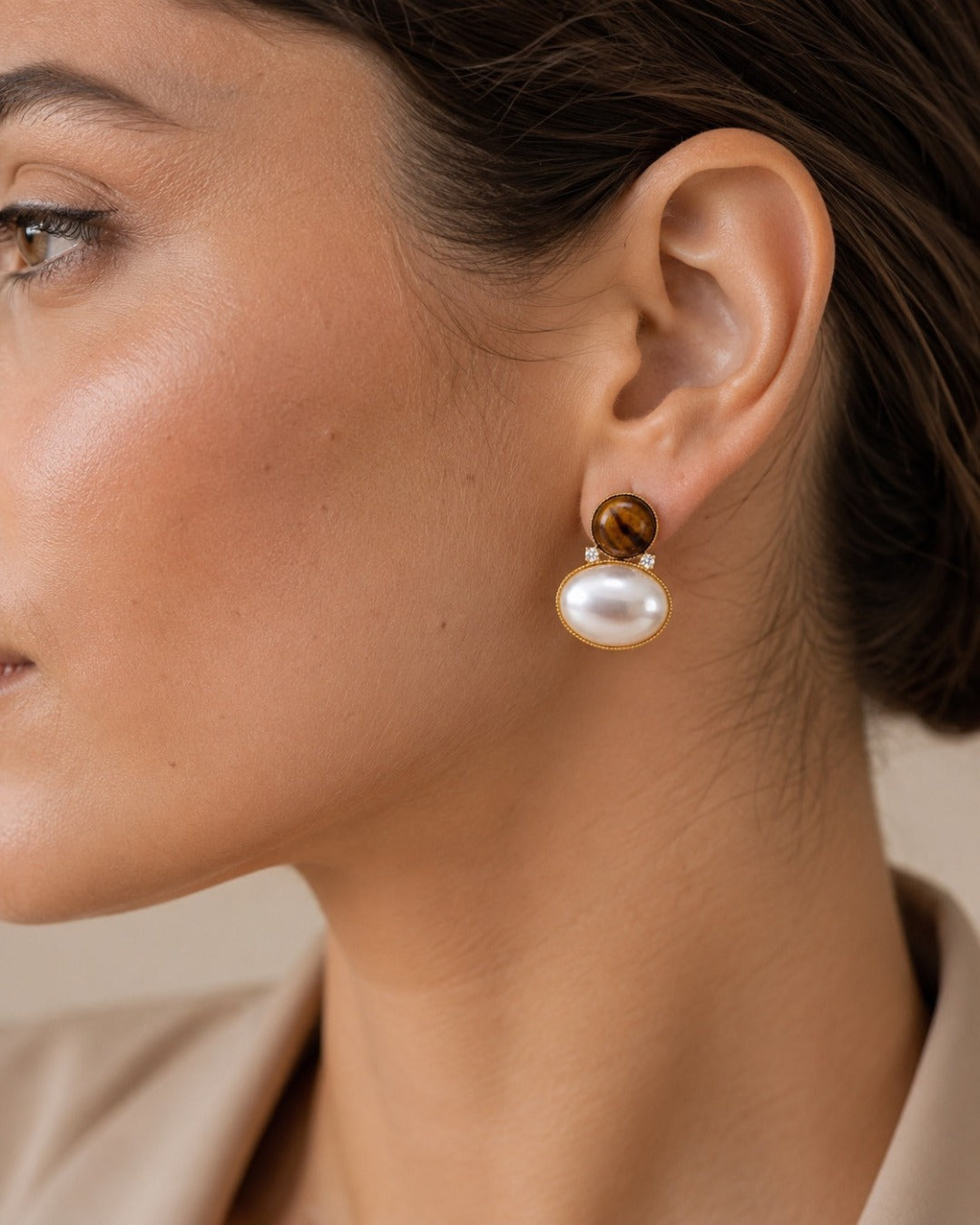 A close-up of a woman's ear wearing a drop earring with a round white pearl and a small gold-toned tiger's eye accent.