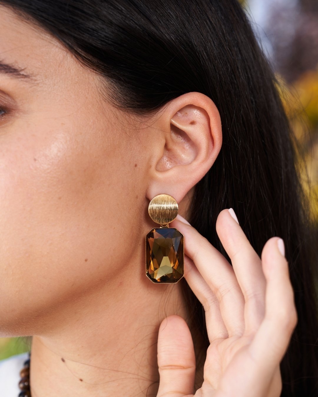 A close-up of a woman wearing a drop earring with a textured gold-tone top and a large faceted amber-brown rectangular drop, while touching the earring with her finger.
