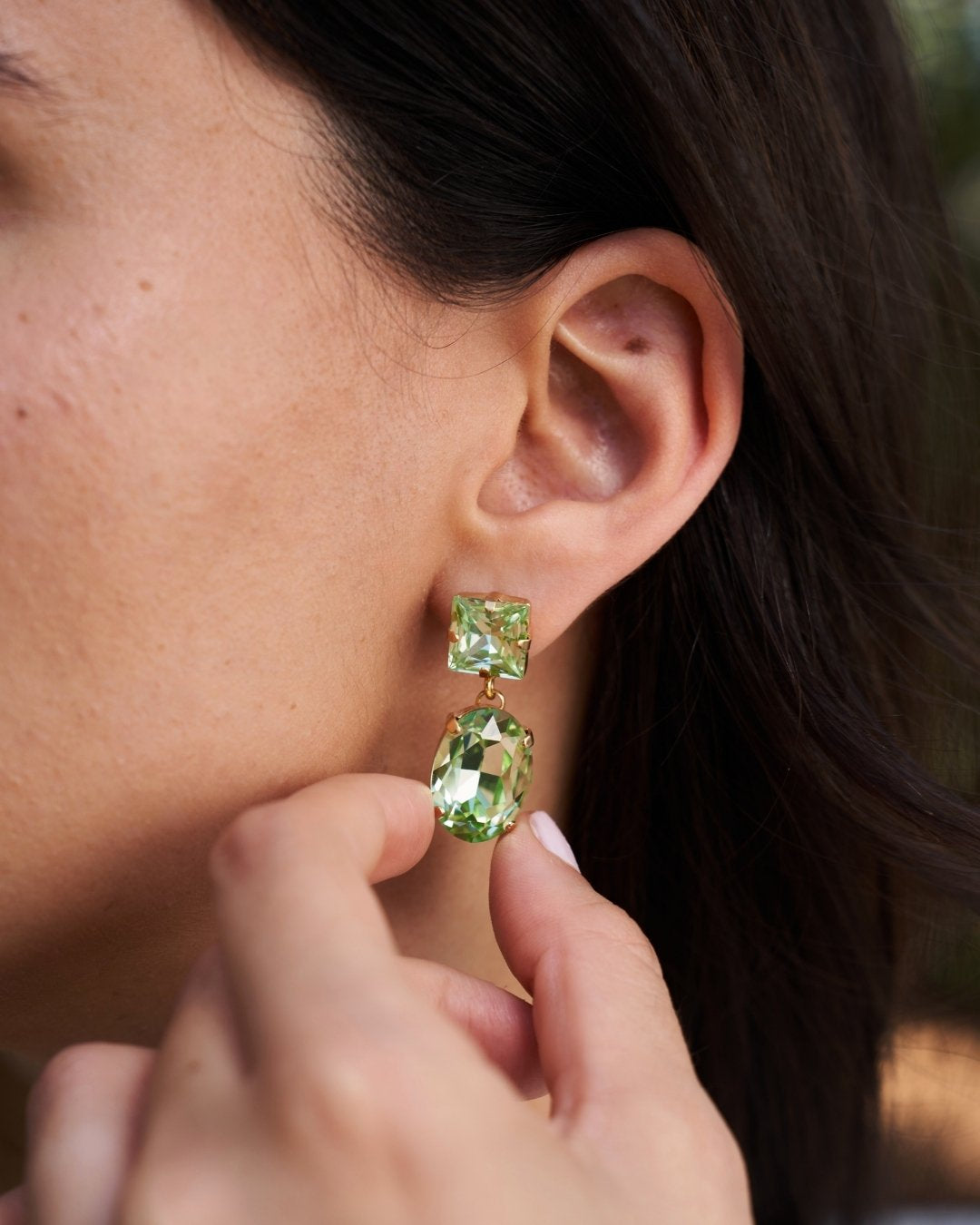 A close-up of a person wearing a drop earring with two green faceted gemstones in a gold-tone setting, with their fingers near the earring.
