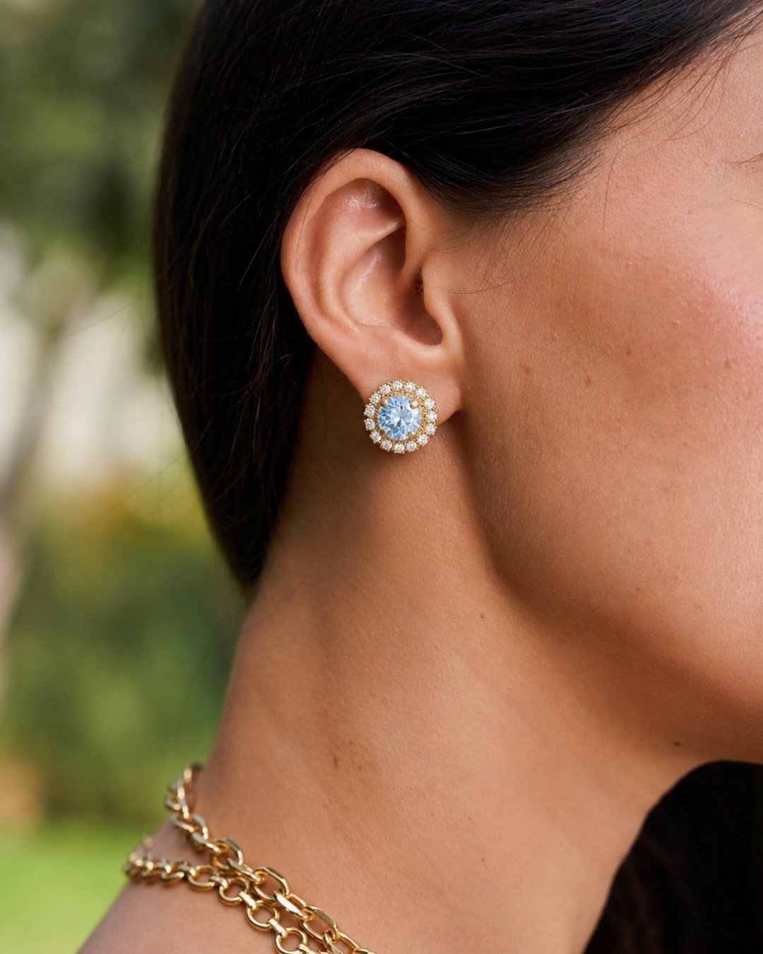 A close-up side profile of a woman wearing a gold stud earring with a central light blue gemstone surrounded by a halo of small clear stones. She is also wearing a gold chain necklace, with a blurred green outdoor background.