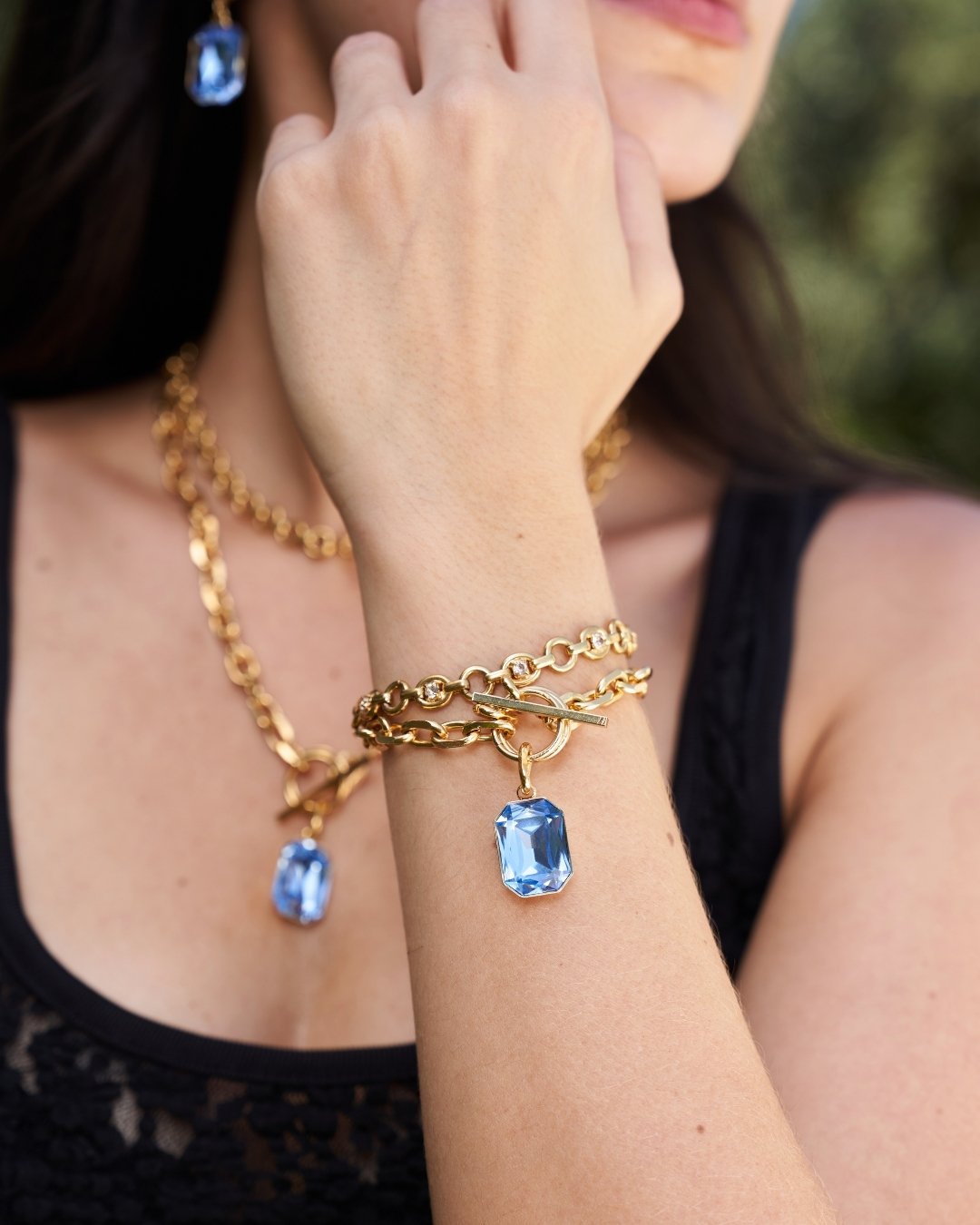 A close-up lifestyle shot of a model wearing a gold chain bracelet with a rectangular blue gemstone pendant, along with matching gold necklaces and earrings.
