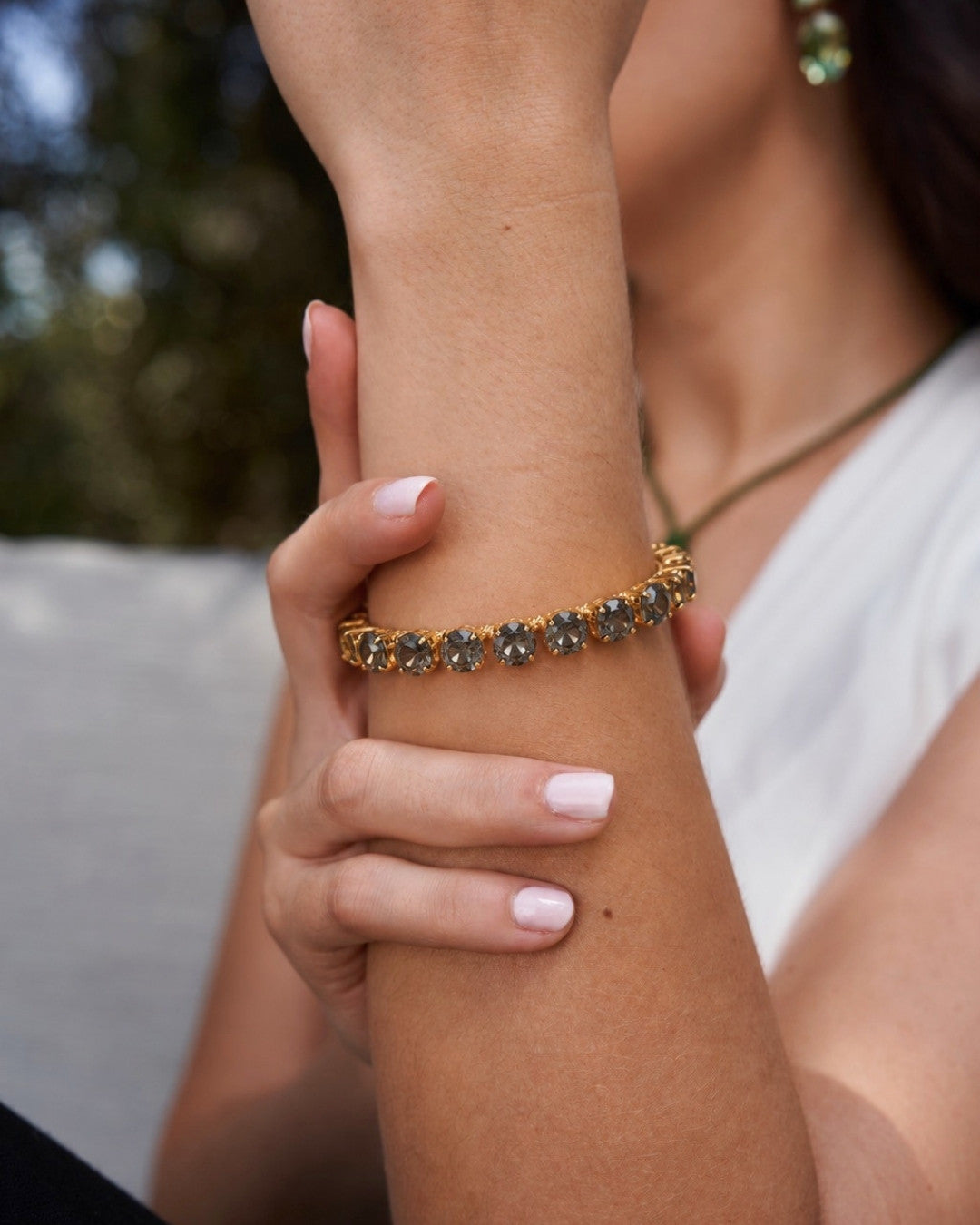 A close-up of a woman's wrist wearing a gold-toned bracelet with small dark gemstones, with her hand resting on her forearm. She is wearing a white top, and the background is softly blurred.