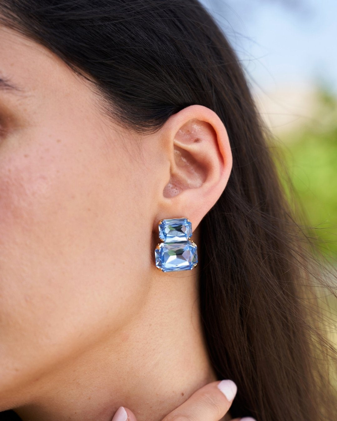 A close-up of a model's ear wearing a pair of gold-toned stud earrings with stacked rectangular light blue gemstones.