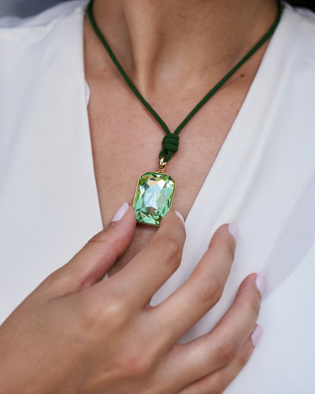 A close-up of a person wearing a green cord necklace with a faceted light green rectangular pendant, with one hand resting near the necklace.