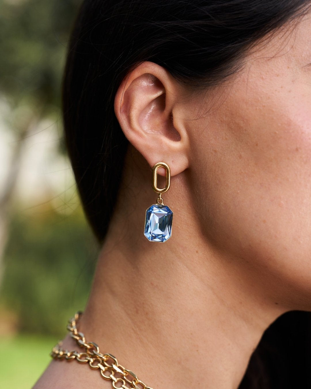 A close-up side profile of a woman wearing a gold drop earring with a rectangular light blue gemstone, along with a gold chain necklace, against a blurred outdoor background.