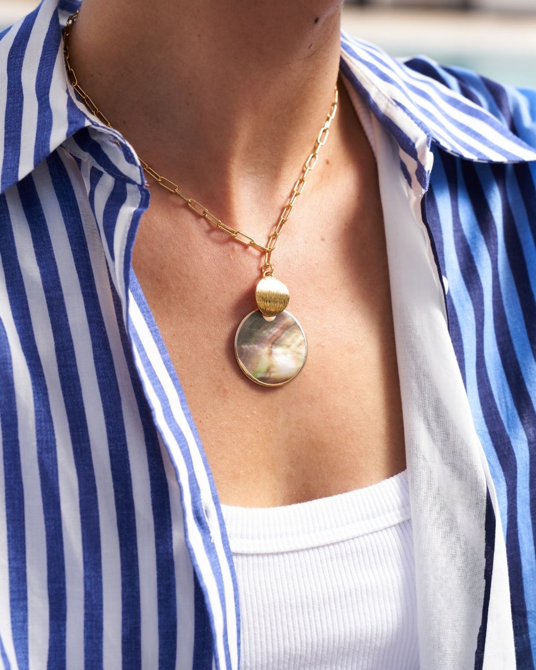 A gold chain necklace with a round mother-of-pearl pendant and a small gold bead accent, worn by a person in a blue and white striped shirt over a white top.