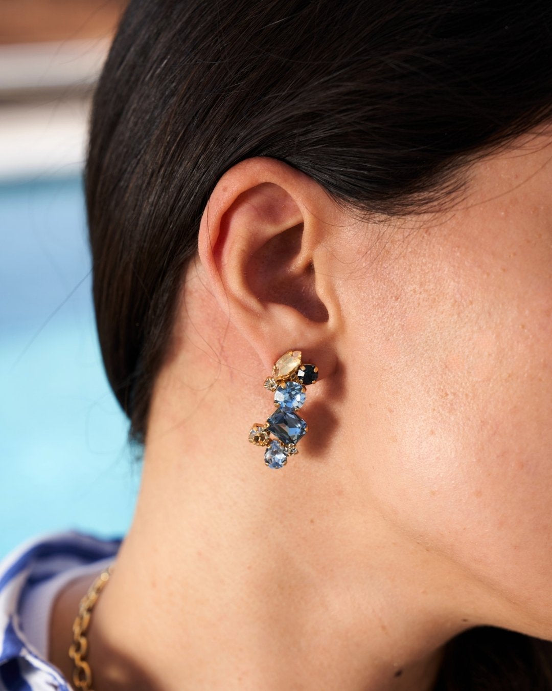 A close-up side profile of a person wearing a dangling earring with light blue stones and gold-toned metal, against a softly blurred background.