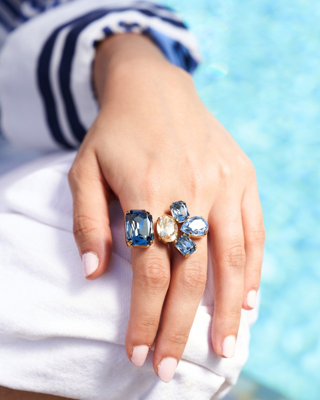 A close-up of a hand wearing a statement ring with blue and clear gemstones in gold settings, resting on white fabric with a blurred blue background.