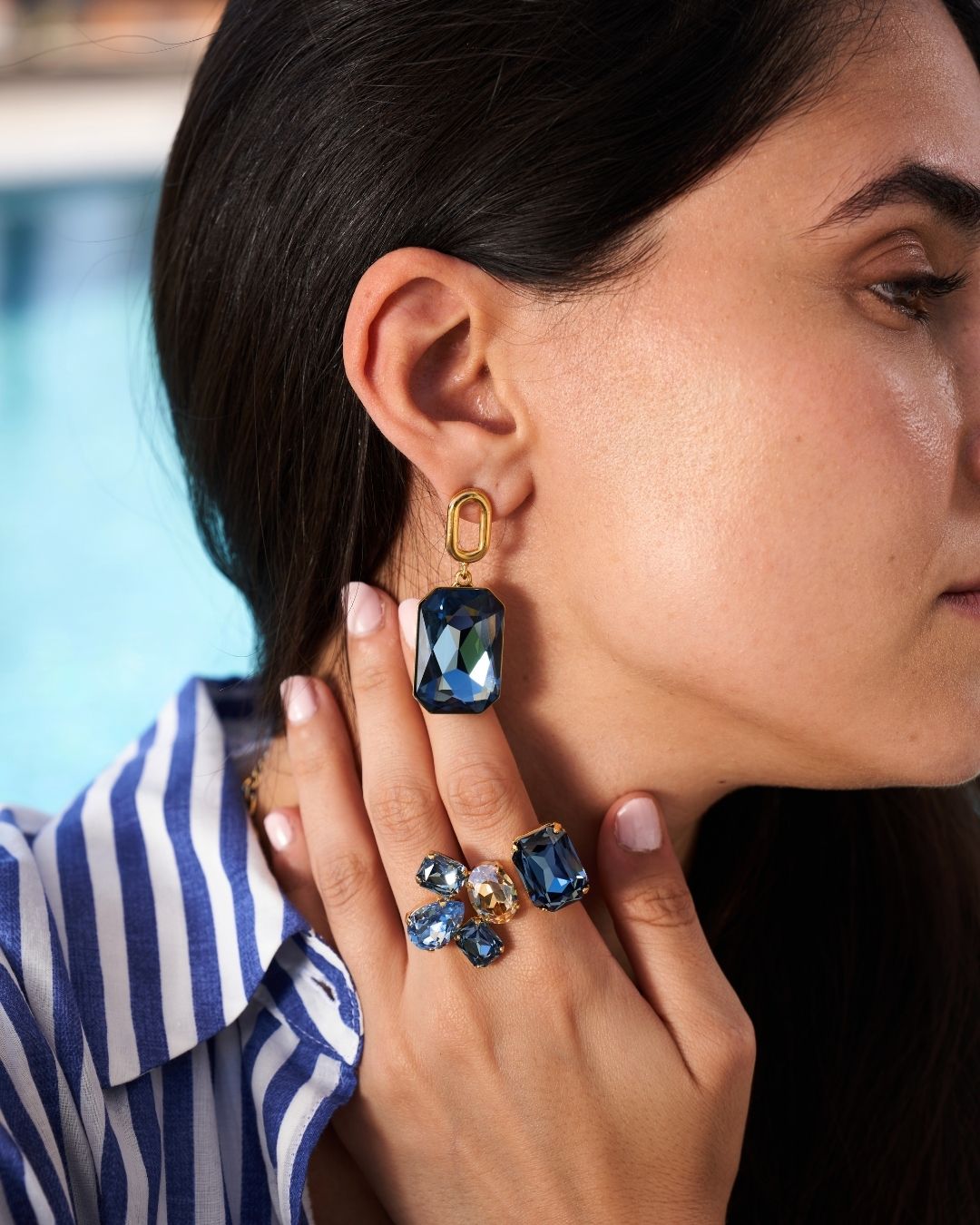 A close-up lifestyle shot of a woman wearing a gold drop earring with a rectangular blue faceted gemstone, along with matching blue gemstone rings on her fingers.