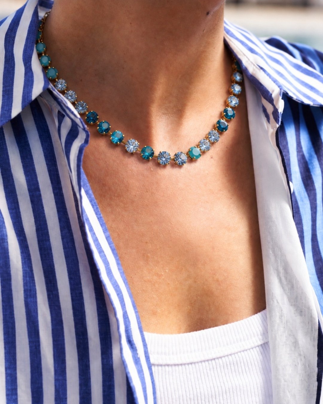 A close-up of a person wearing a delicate necklace with alternating turquoise-blue and clear faceted stones, styled with a blue and white striped shirt.