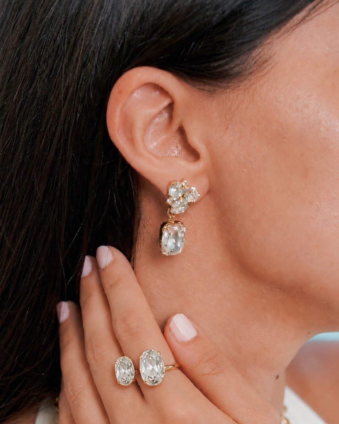 Close-up of a woman wearing a gold drop earring with clear gemstones and matching oval gemstone rings on her fingers.