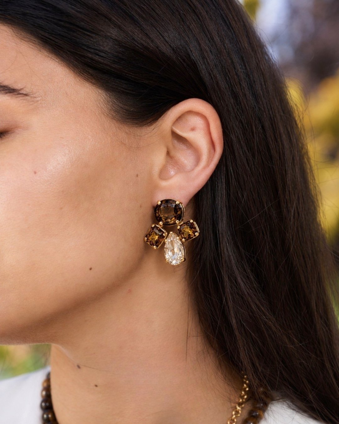A close-up side profile of a woman wearing a gold-toned drop earring with dark brown and clear teardrop-shaped crystals.