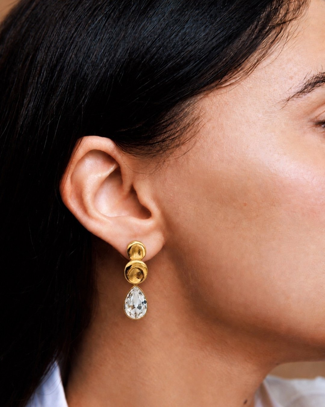 Close-up of a person wearing gold earrings with a clear gemstone.