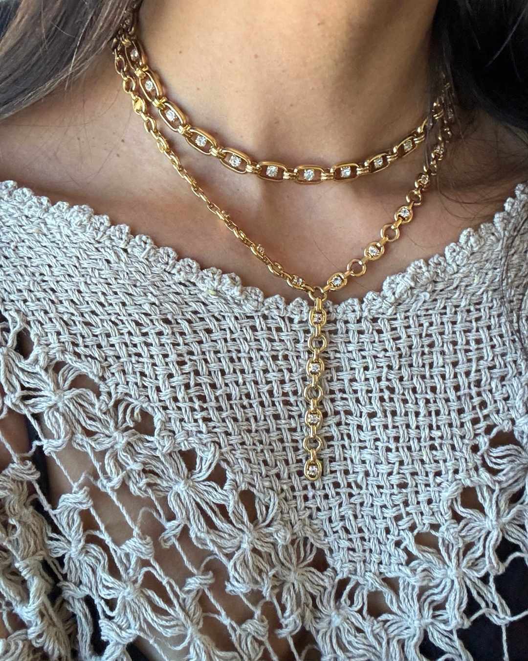 A close-up of a person wearing layered gold necklaces, including a choker and a longer Y-shaped pendant necklace, paired with a white crochet top.