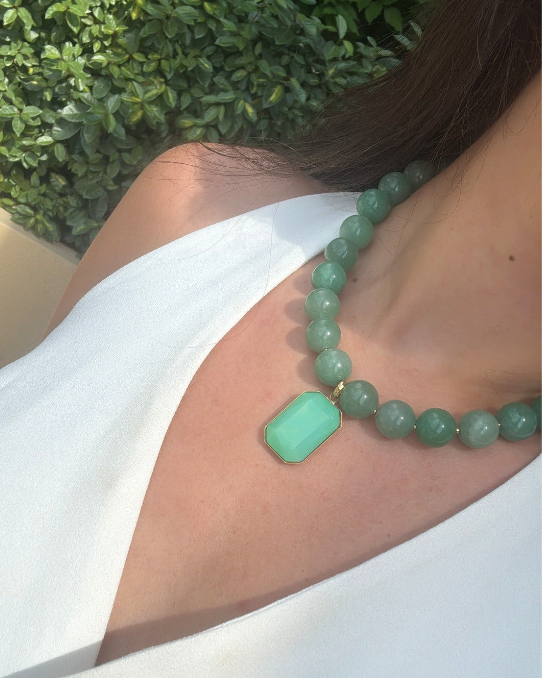 A close-up of a woman wearing a green beaded necklace with a rectangular mint-green pendant, styled with a white top against a background of green foliage.