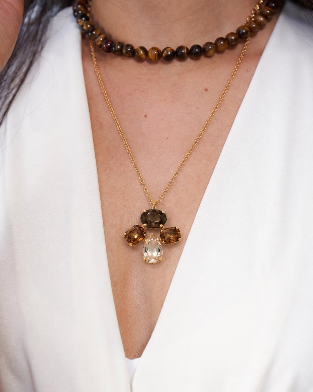 A close-up of a person wearing layered necklaces: a gold chain pendant necklace with a cross-shaped gemstone design featuring a clear teardrop and brown smoky stones, and a brown beaded choker.