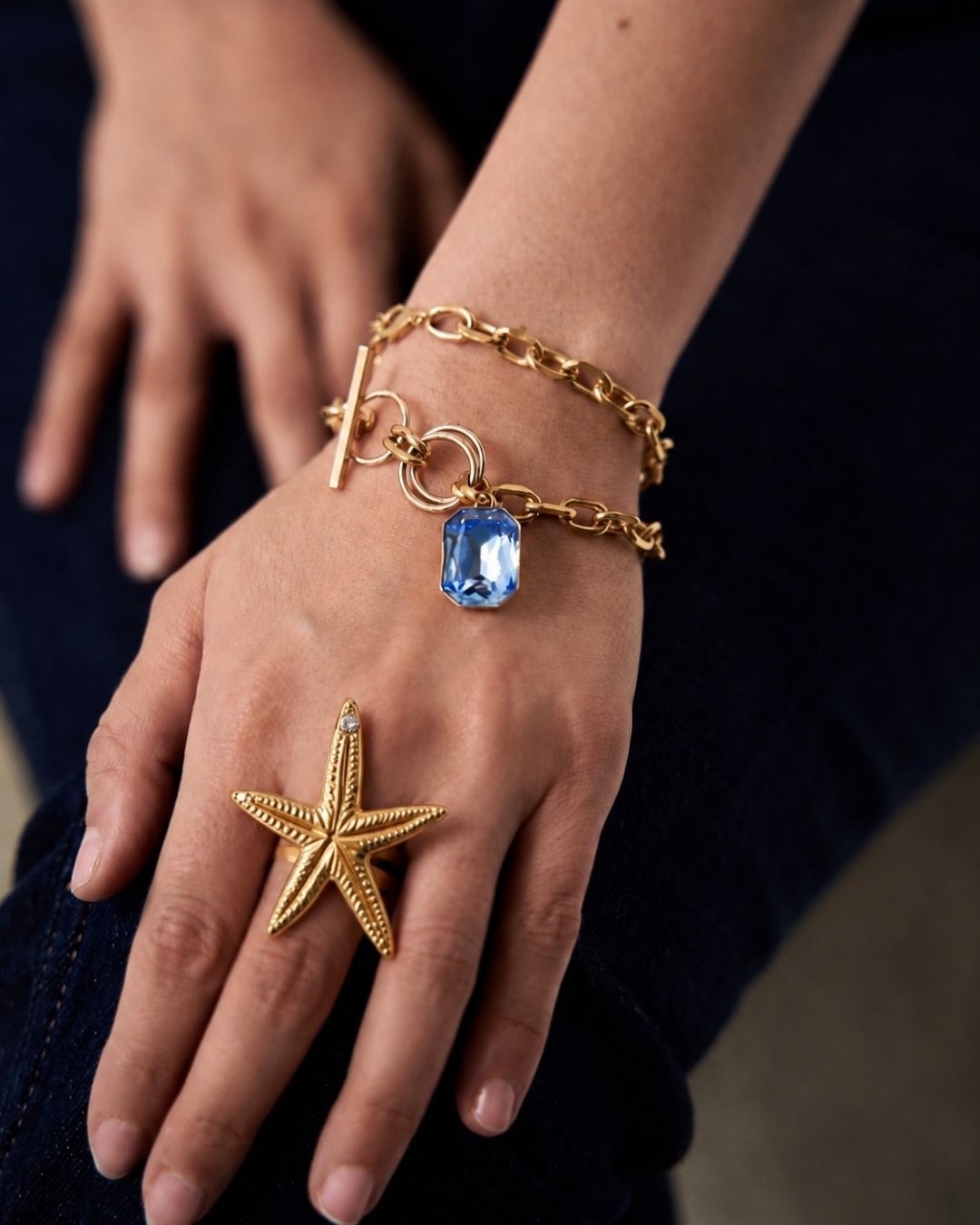 Hand wearing a gold bracelet with a blue gemstone and a starfish ring on a dark background