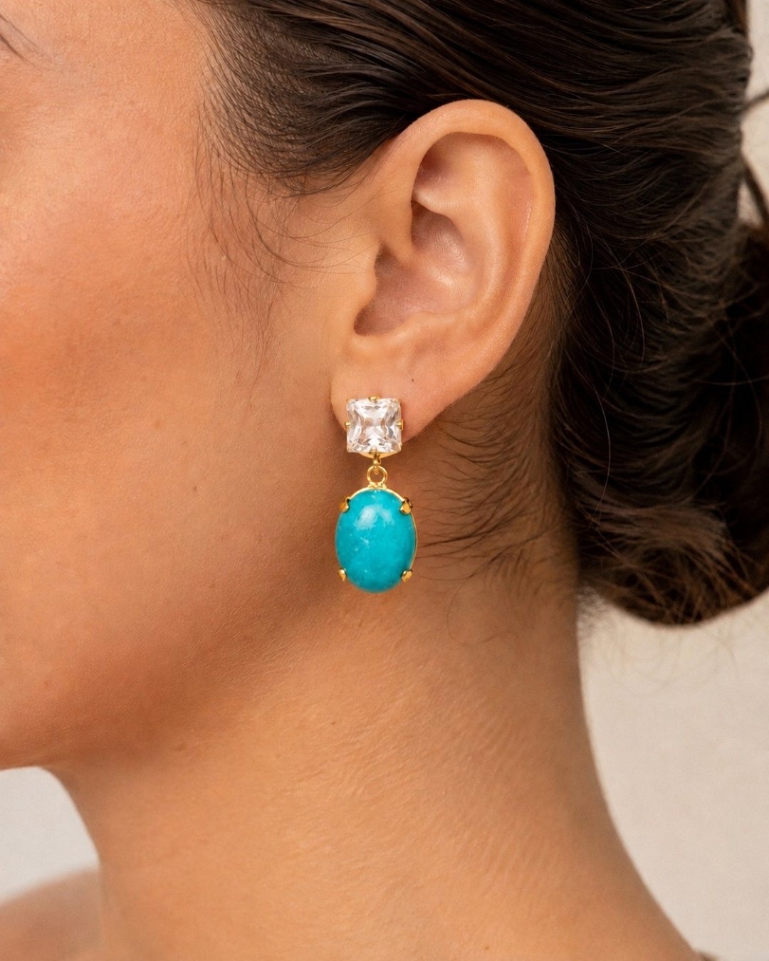 A close-up of a model's ear wearing a gold drop earring with a clear square stud and a dangling oval turquoise stone.