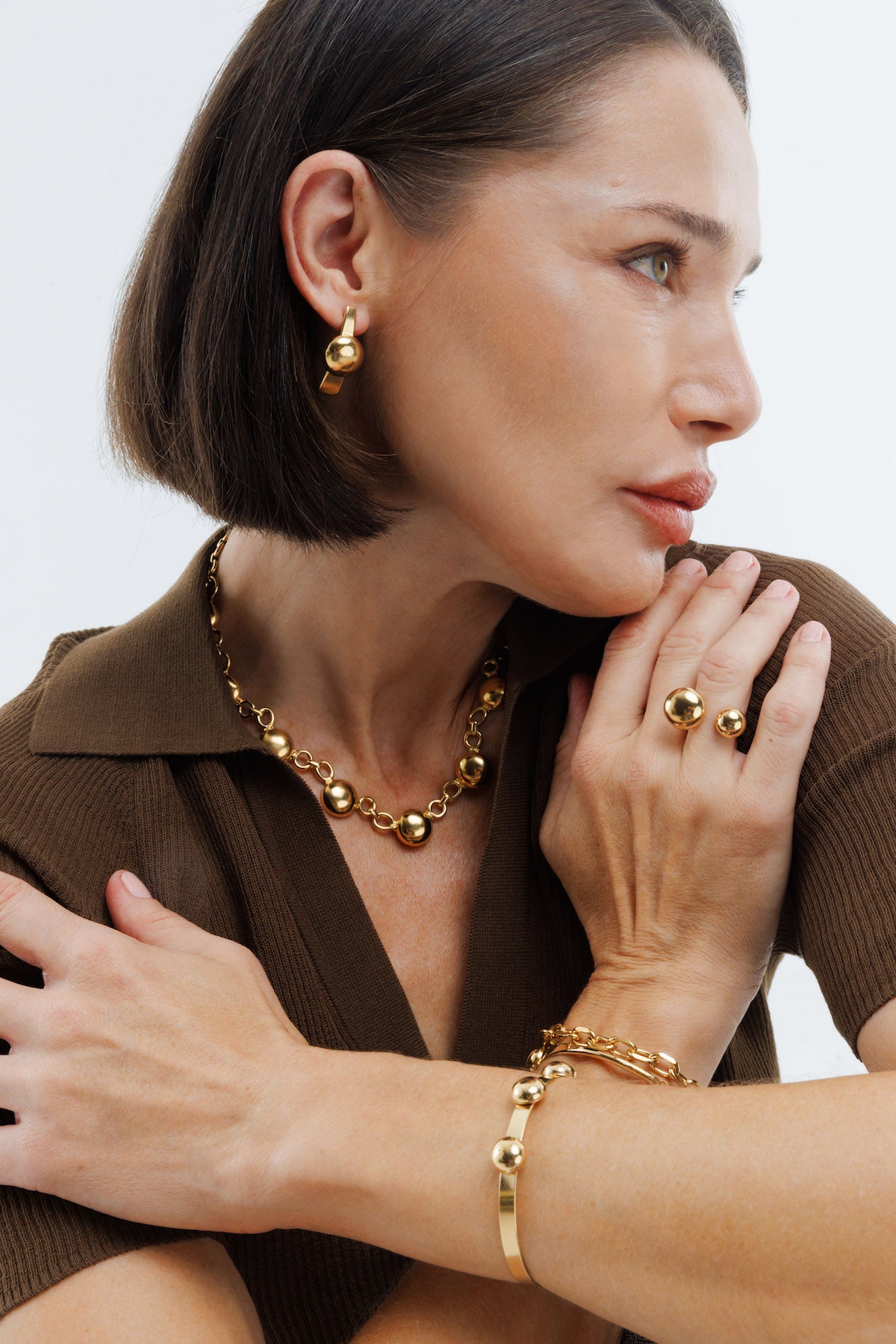 Woman wearing gold jewelry including earrings, necklace, ring, and bracelet against a neutral background