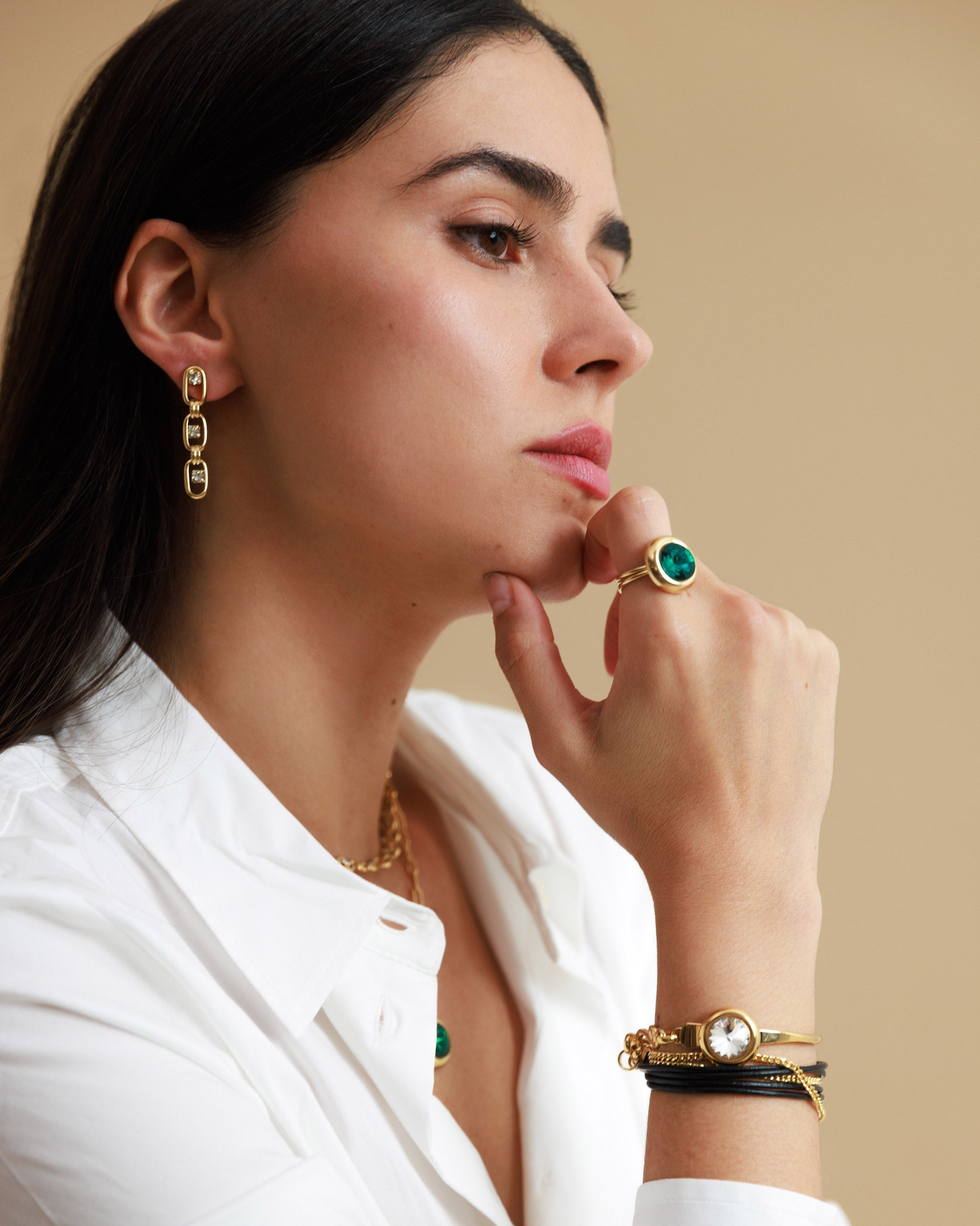 Woman wearing gold jewelry including earrings, ring, and bracelets on a beige background