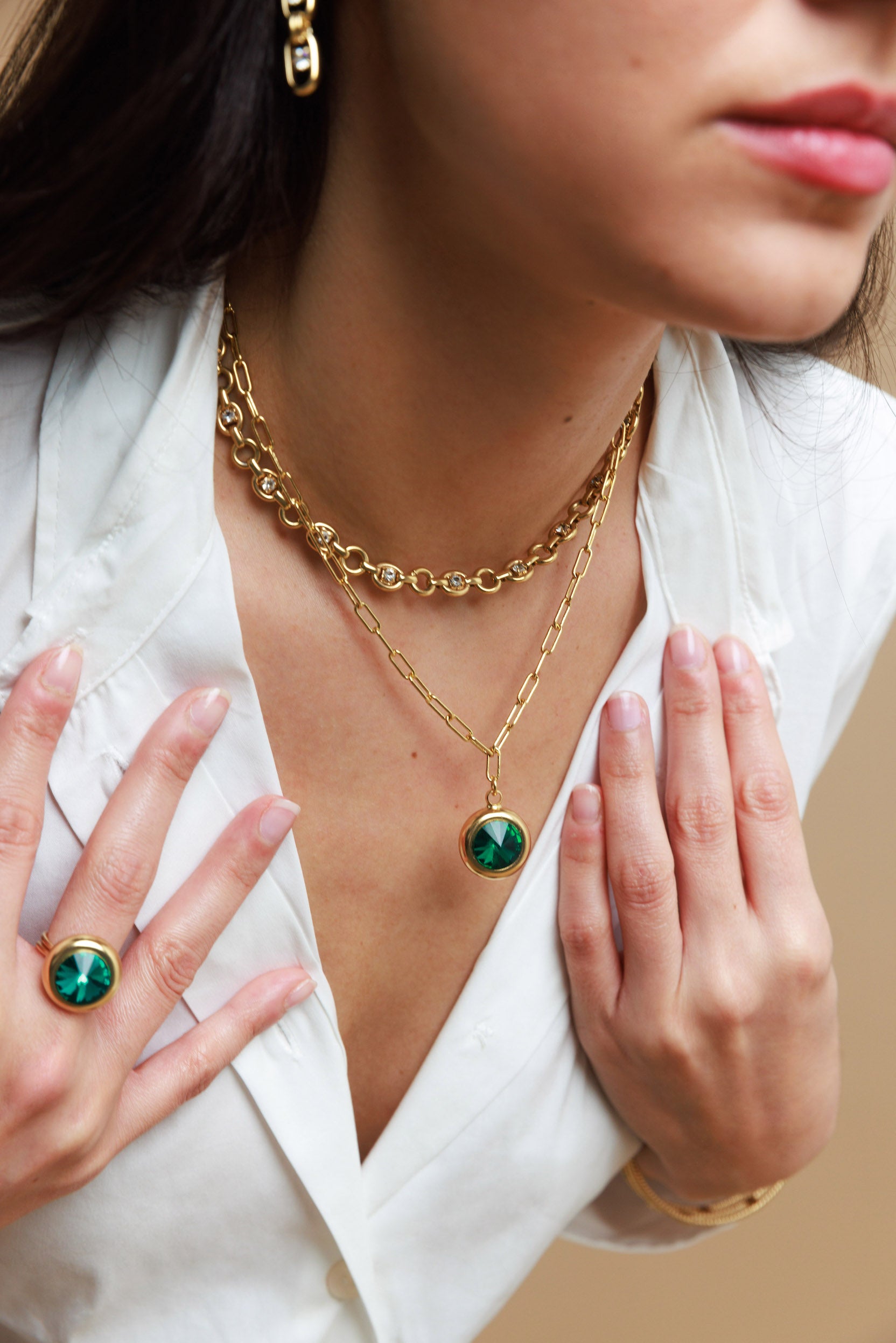 Woman wearing gold necklaces and rings with green gemstones on a neutral background