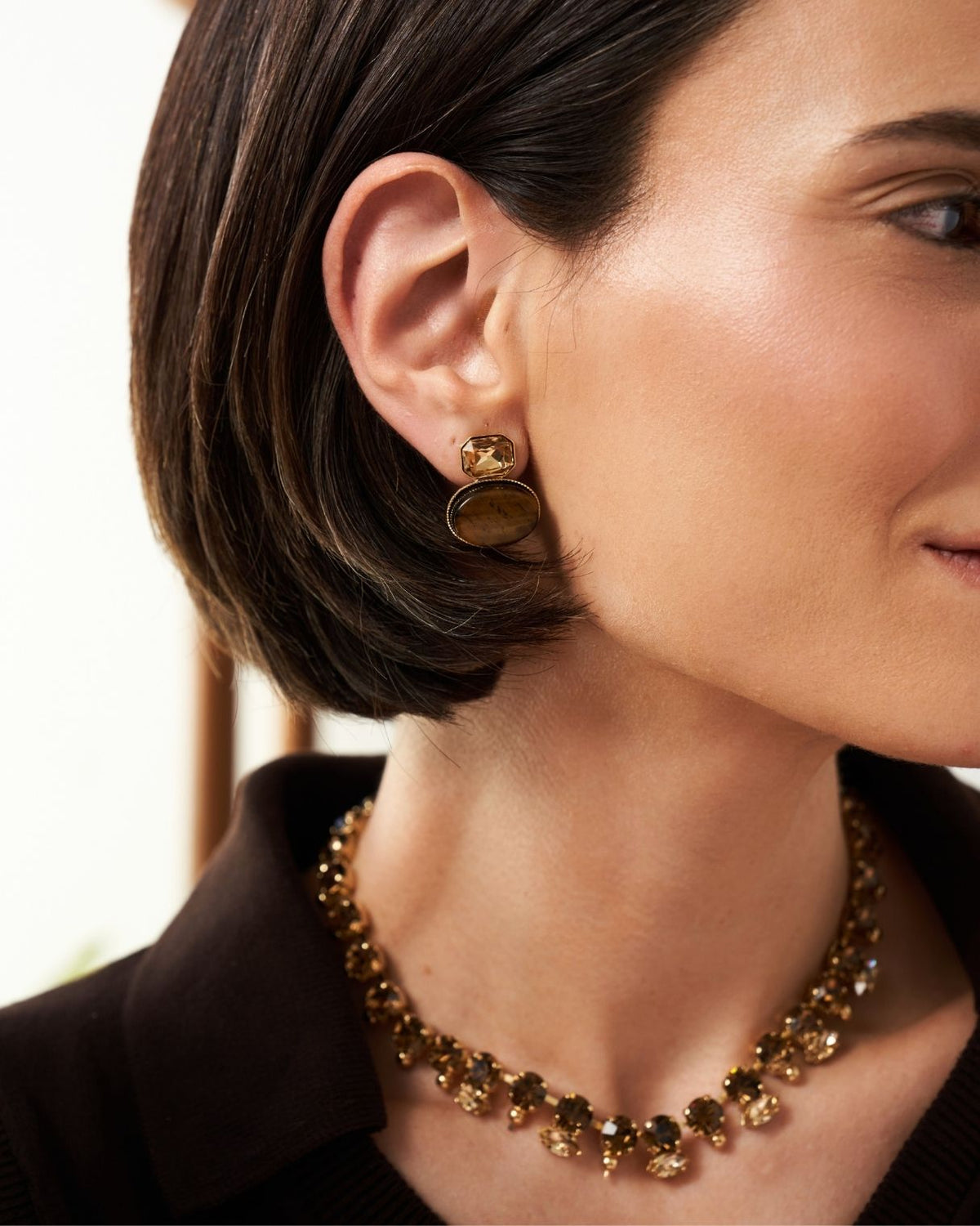 A close-up of a woman wearing a gold-toned statement necklace and matching drop earrings with dark brown and amber stone details.
