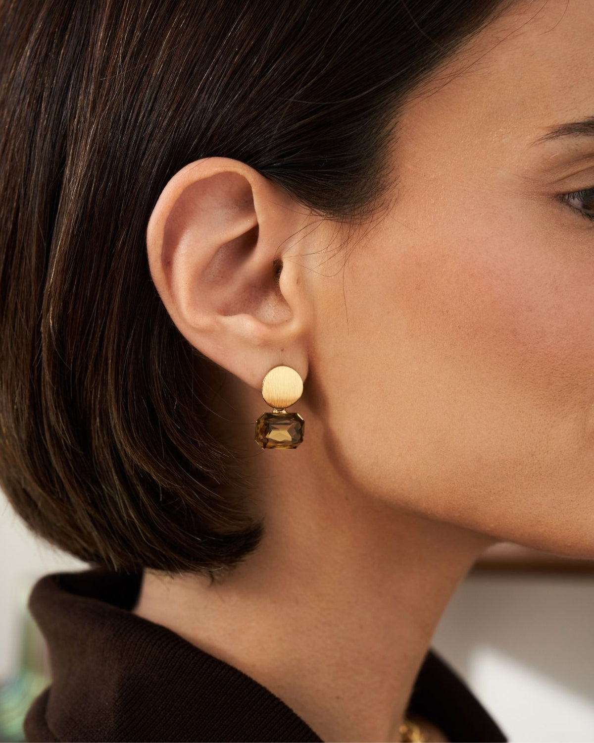 A close-up of a model's ear wearing a double-drop statement earring. The earring features a round, pale cream-colored top disc and a rectangular, faceted dark brown gemstone drop below it.