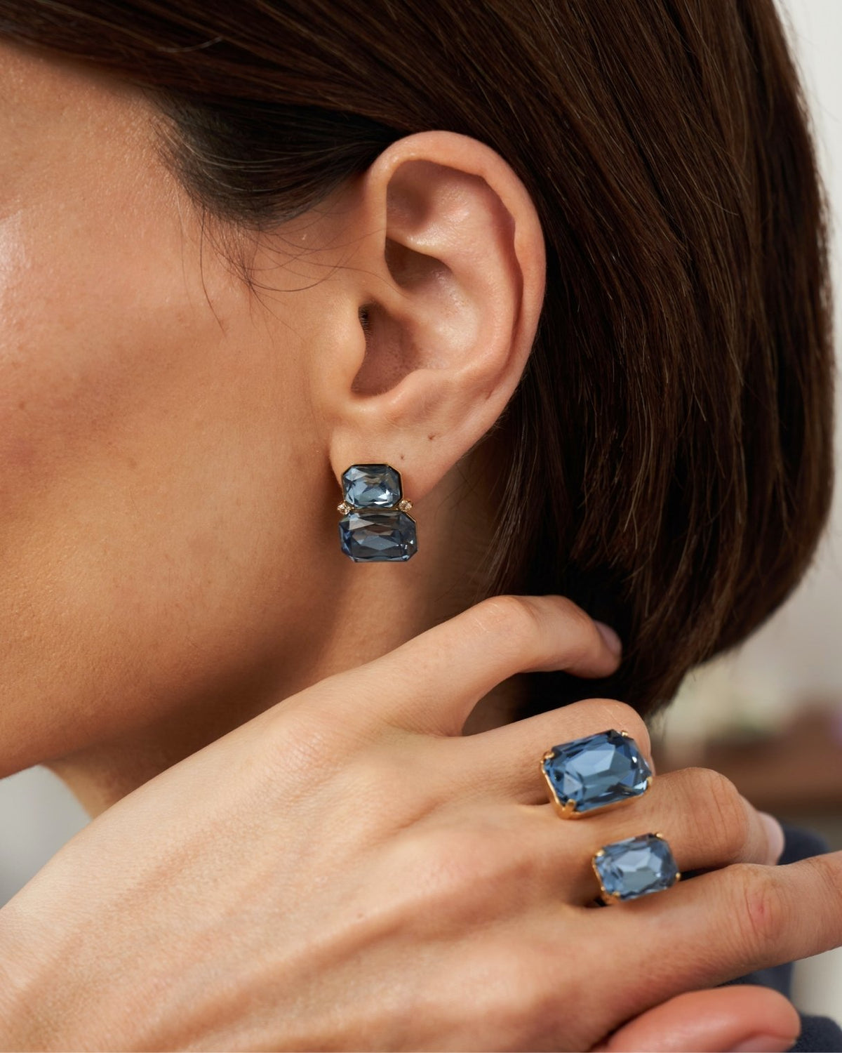 A close-up lifestyle shot of a model wearing matching jewelry, featuring a pair of rectangular blue gemstone stud earrings and two matching blue gemstone rings with gold settings.