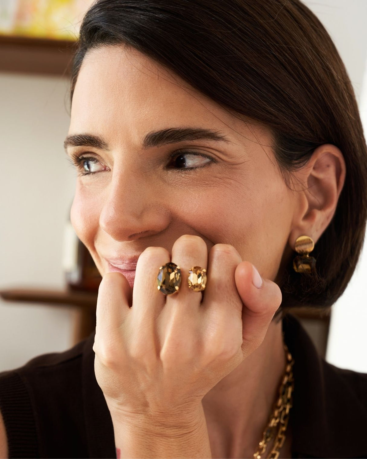A close-up lifestyle shot of a woman with dark brown hair, wearing gold jewelry including a chunky gold ring with two dark oval gemstones on her finger, a matching gold earring, and a gold chain necklace.