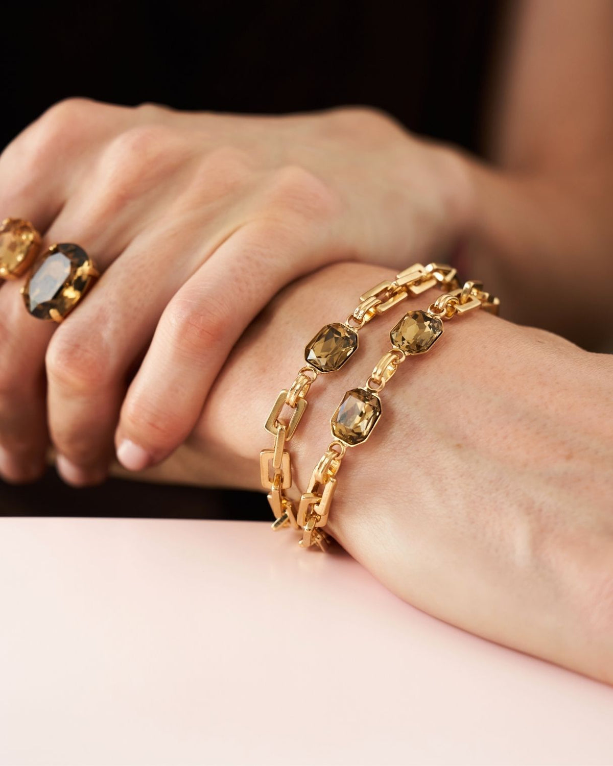 A close-up of a person's hands wearing a gold chain bracelet with rectangular faceted clear stones, alongside a matching gold ring with a dark stone.