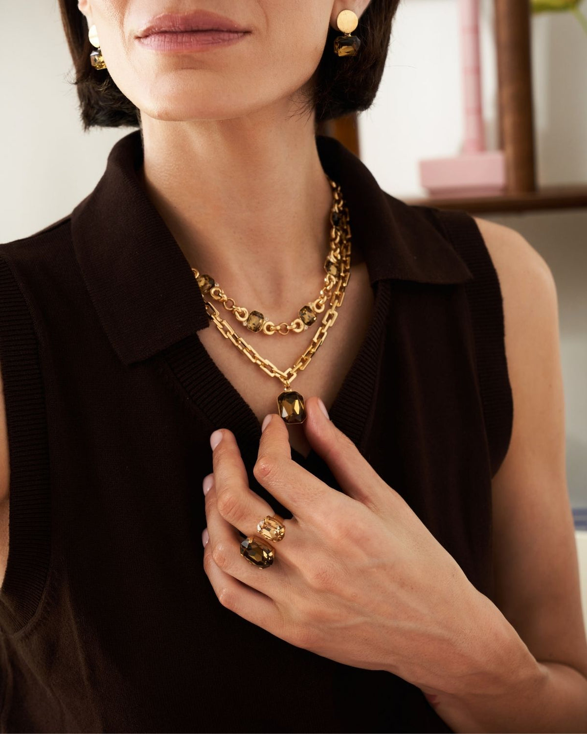 A close-up lifestyle shot of a model wearing a layered gold chain necklace with a large oval pendant, matching gold rings, and gold hoop earrings, styled with a dark brown sleeveless top.