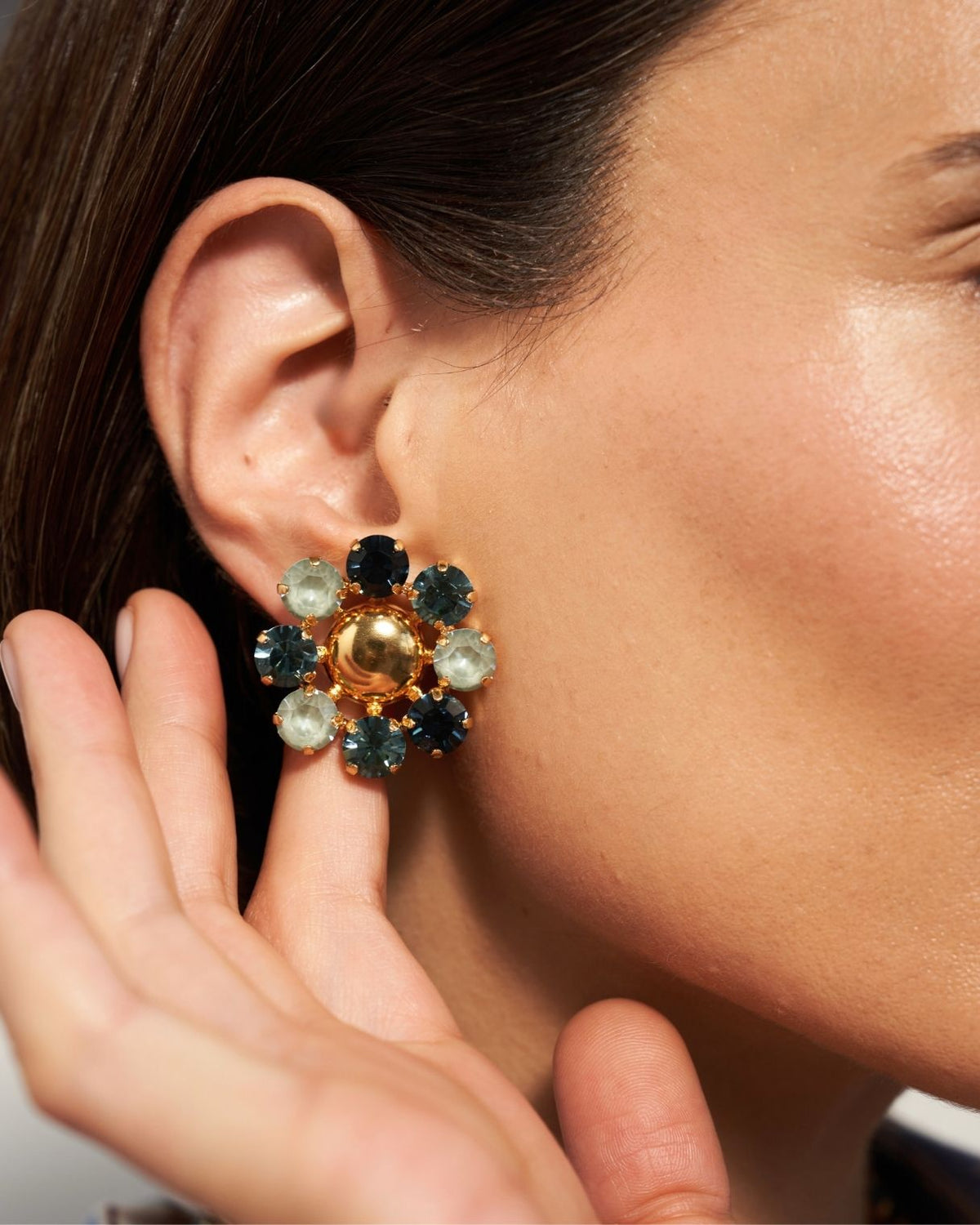 A close-up of a model's ear wearing a gold flower-shaped earring with dark blue, light blue, and white gemstones.