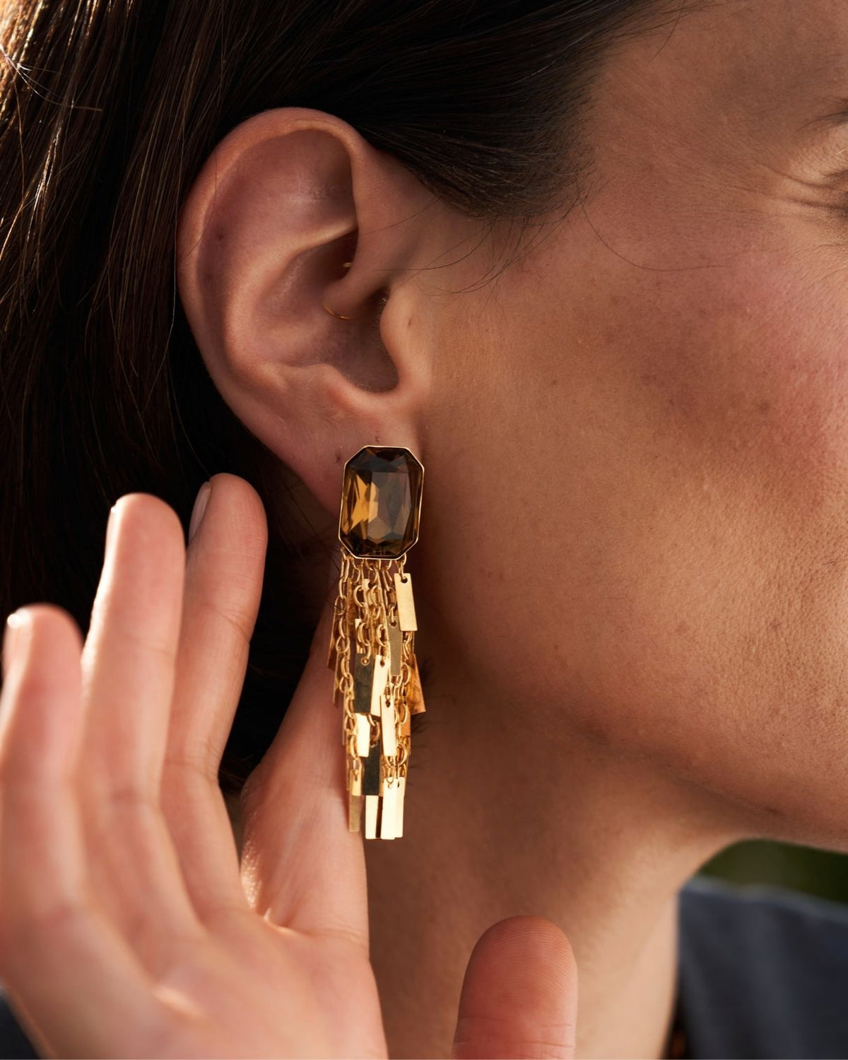 A close-up of a model's ear wearing a gold statement earring with a dark smoky rectangular stone and dangling layered gold chains.