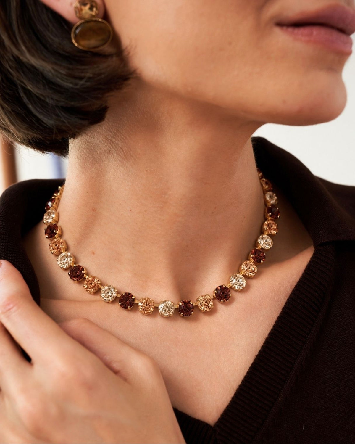 A close-up lifestyle shot of a model wearing a delicate gold necklace with alternating round gold and brown faceted gemstone clusters, paired with a dark brown knit top.