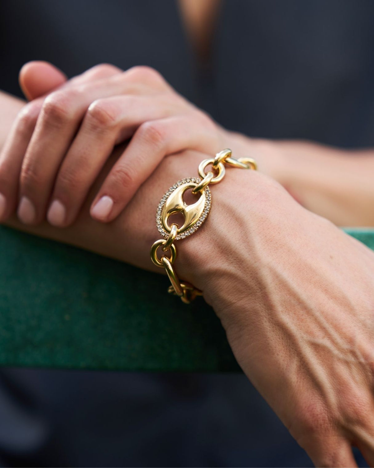 A close-up of a person's wrist wearing a gold chain bracelet with a prominent oval link featuring a pavé-set stone center.