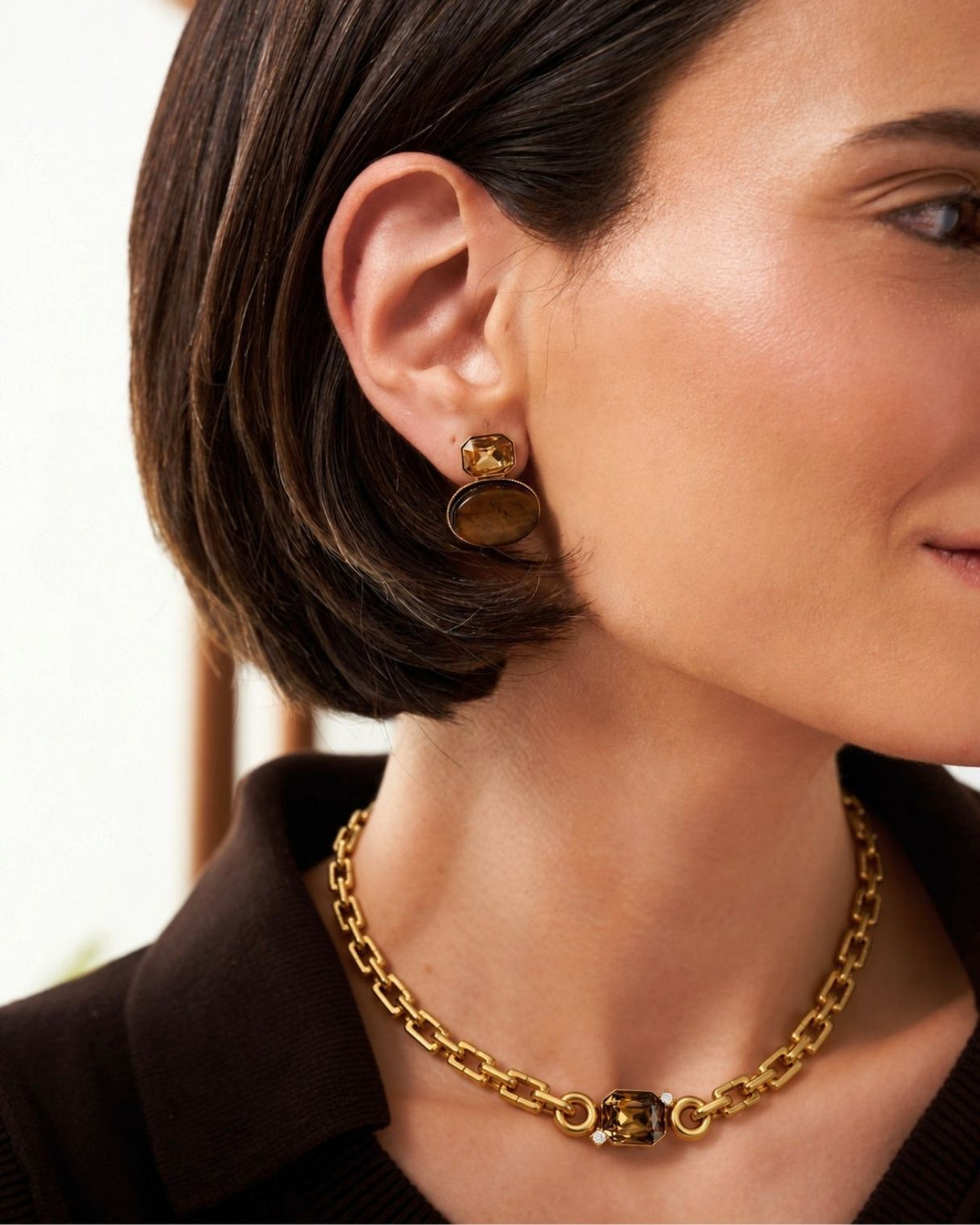 Close-up of a woman wearing a gold chain necklace with a central square charm and matching gold drop earrings, paired with a dark brown top.