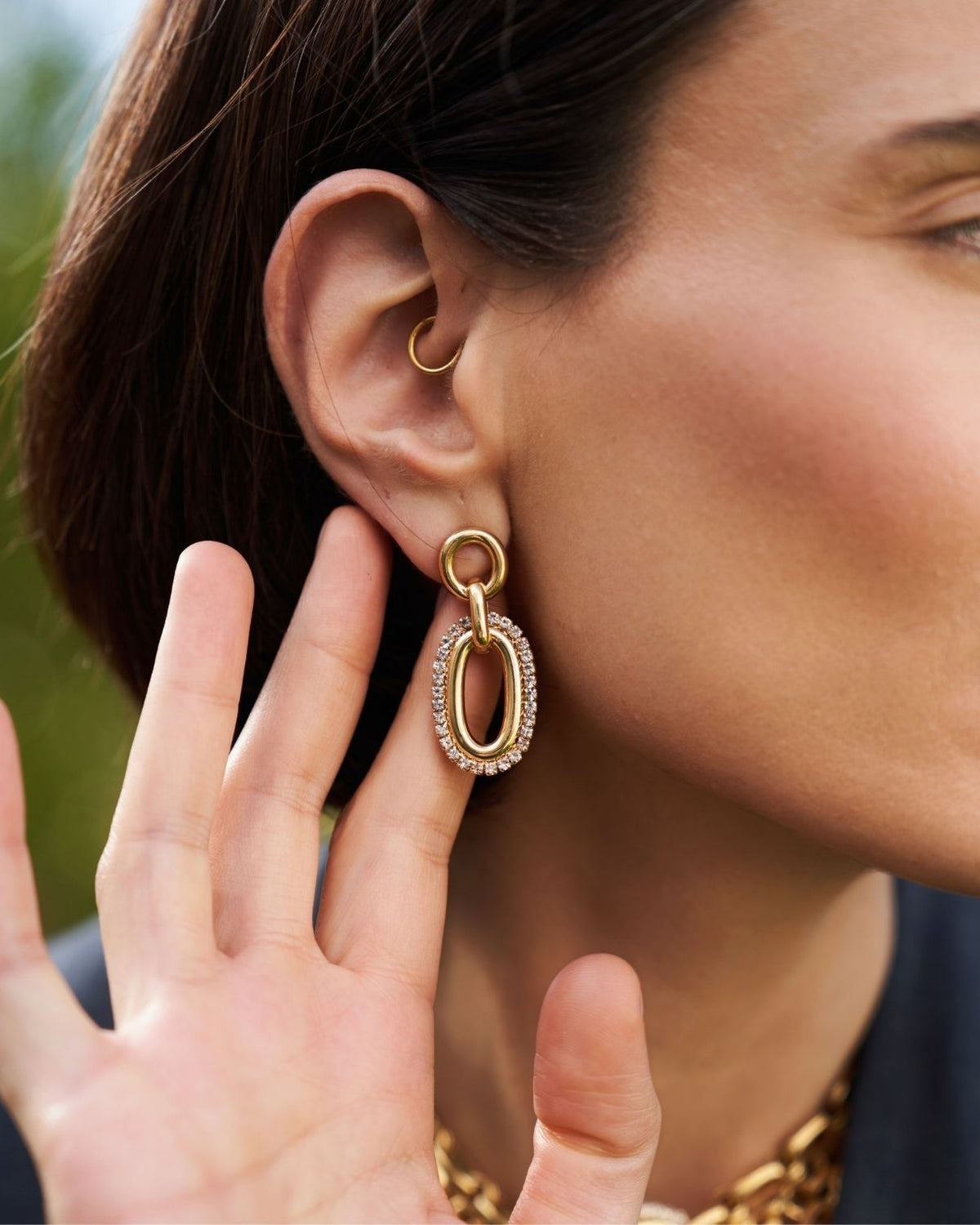 A close-up of a woman's ear wearing a gold hoop earring in the lobe and a larger gold drop earring with a clear crystal border, against a softly blurred outdoor background.