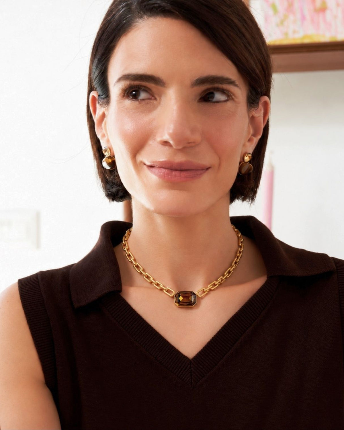 A woman with short dark hair wearing a gold chain necklace with a dark brown oval centerpiece and matching gold drop earrings, paired with a dark brown sleeveless collared top.