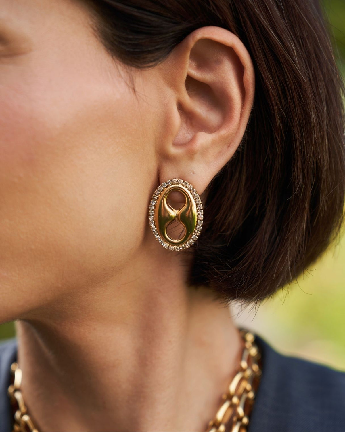 A close-up of a model's ear wearing a gold oval stud earring with a central abstract cutout and a border of small clear stones.