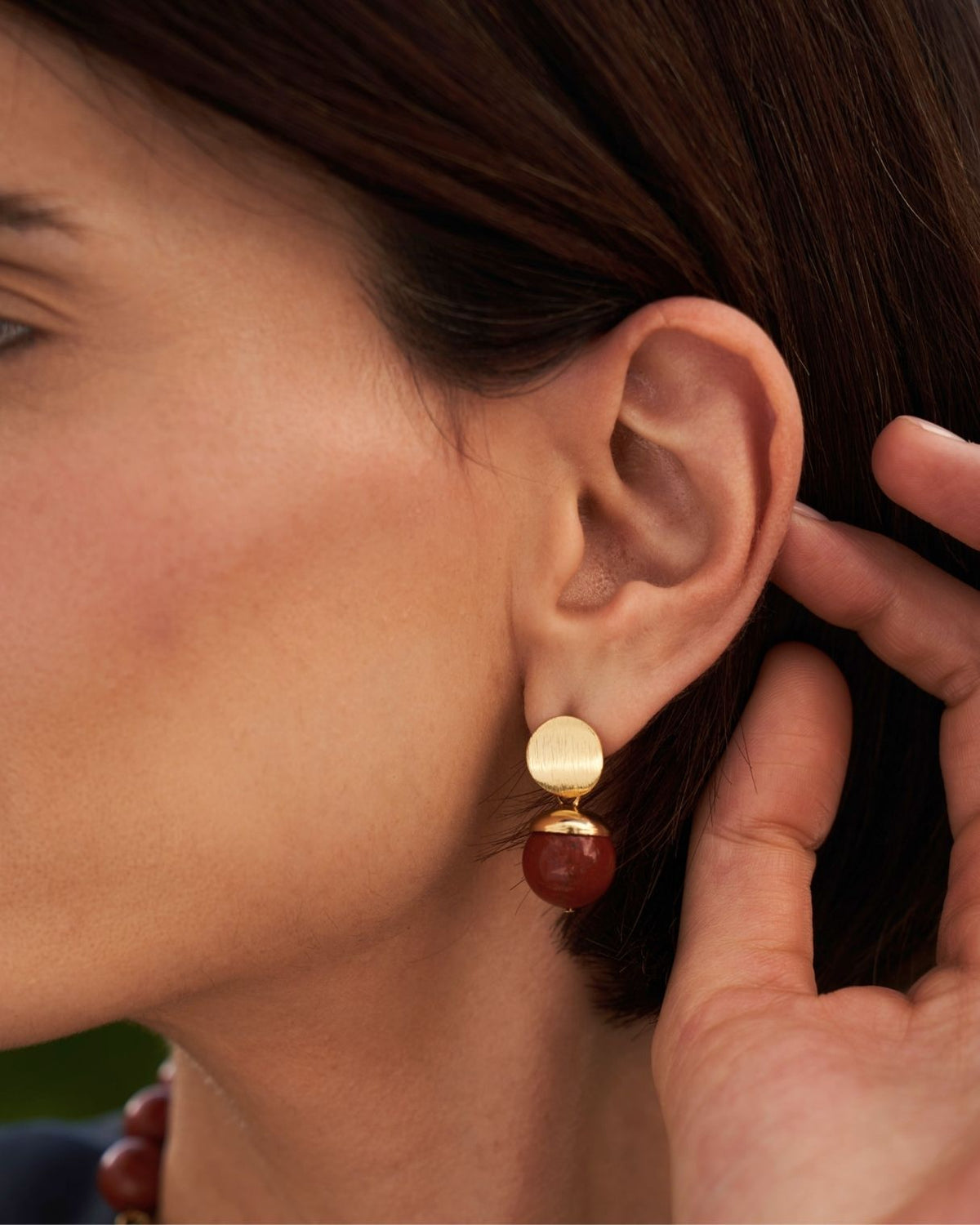A close-up of a woman's ear wearing a double drop earring with a gold-toned flat top disc and a round reddish-brown bead drop.