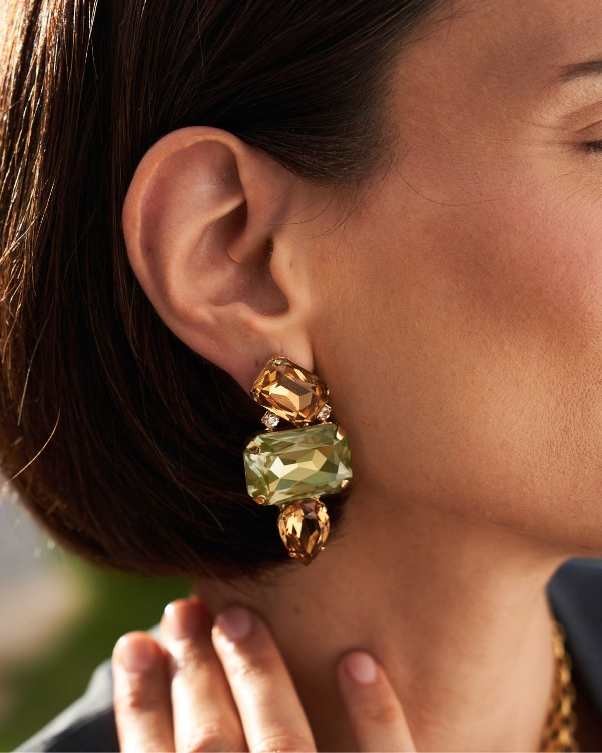 A close-up of a model wearing a large gold-toned statement earring with a rectangular pale green gemstone and smaller gold stones above and below it.